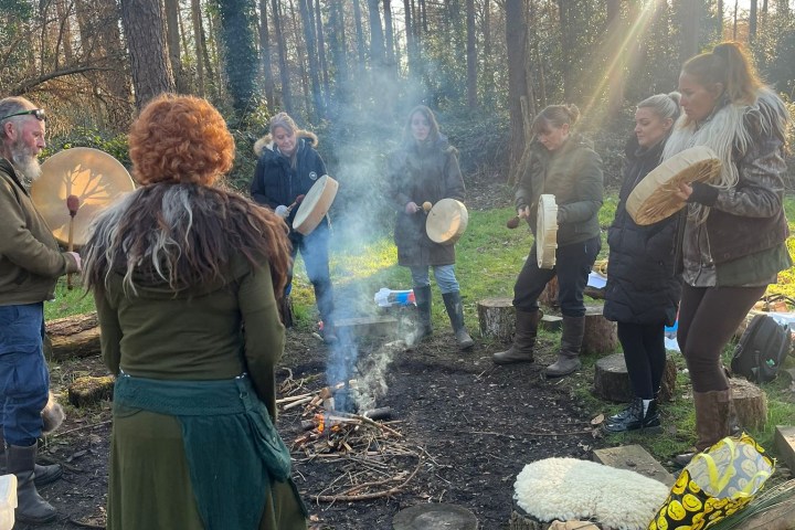 People in a forest gather around a small campfire with drums.